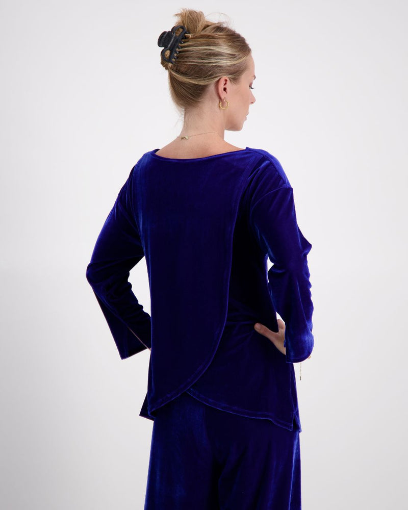 A woman is shown from the back on a 3/4 angle, wearing a cobalt blue velvet boat neck top. Her hair is held up with a clip, and she wears small hoop earrings. The back of the top features a leaf-back design.