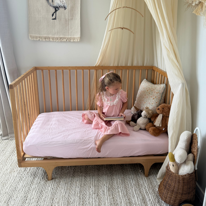 Child in a pink dress sitting on a wooden bed with stuffed animals in a cozy room.