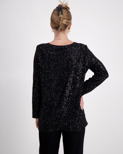 This is a photograph of a woman posing against a white background with her back towards the camera. She has her right hand on her hips. Her blonde hair is styled in an up-do, and she is wearing a black sequinned v-neck top with full length sleeves, paired with black sequin pants. 