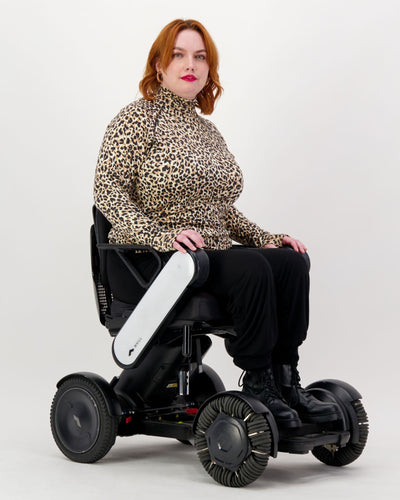 Our seated model with red hair and who is in a wheelchair is wearing a leopard print thermal top with long sleeves, black pants and black boots. Christina Stephens Adaptive Clothing Australia.