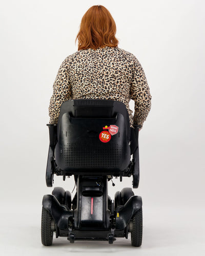 Our seated model with red hair and who is in a wheelchair is wearing a leopard print thermal top with long sleeves, black pants and black boots. Christina Stephens Adaptive Clothing Australia.