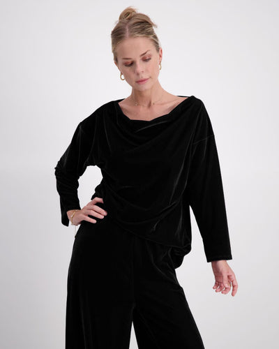 A woman in a midnight black velvet top with draped neckline and matching wide leg pants stands with one hand on her hip, looking downwards against a white backdrop.