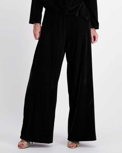 Jessie Wide Leg Velvet Pants in luxurious black velvet, featuring a flowing wide-leg silhouette, side pull tabs, and floor-length design. Elegant evening wear pants shown with metallic sandals against a light gray background.