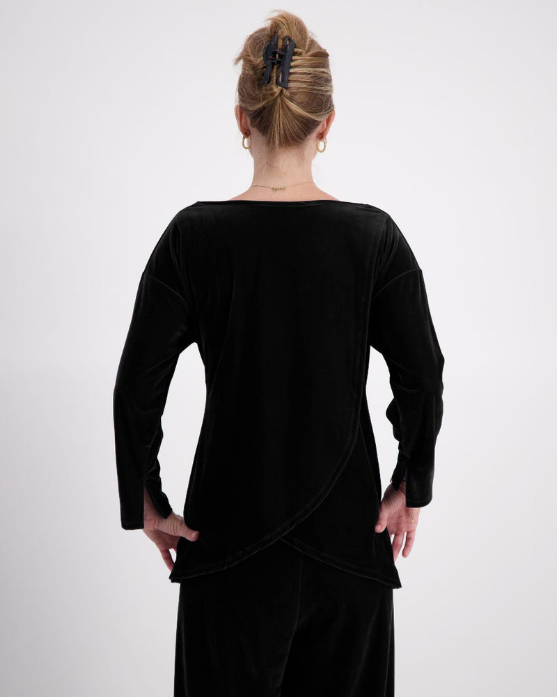 A woman is shown from the back, wearing a midnight black velvet boat neck top. Her hair is held up with a clip, and she wears small hoop earrings. The back of the top features a leaf-back design.