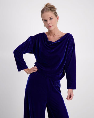  A woman in a cobalt blue velvet top with draped neckline and matching wide leg pants stands with one hand on her hip, looking directly at the camera against a white backdrop.