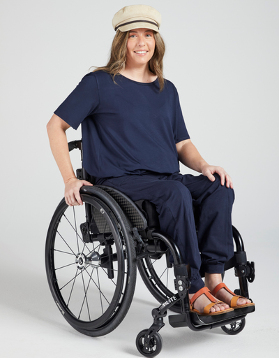 Dark blonde woman in a wheelchair wearing a navy, short sleeve t-shirt, beige cap and navy pants. Orange sandels on her feet. Forward facing and smiling. 