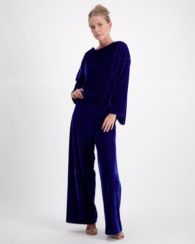 A woman stands with her left leg forward and her hand resting on her upper thigh. She's looking at the camera, wearing cobalt blue velvet wide leg pants and a matching boat neck top. She is in front of a white background and wears open-toed heels.