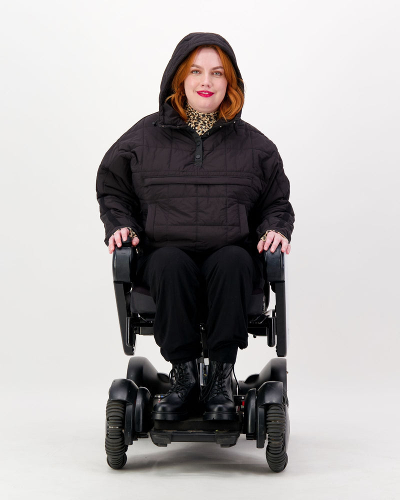 Seated female in a power chair is wearing a black puffer jacket over black pants. Christina Stephens Adaptive Clothing Australia.