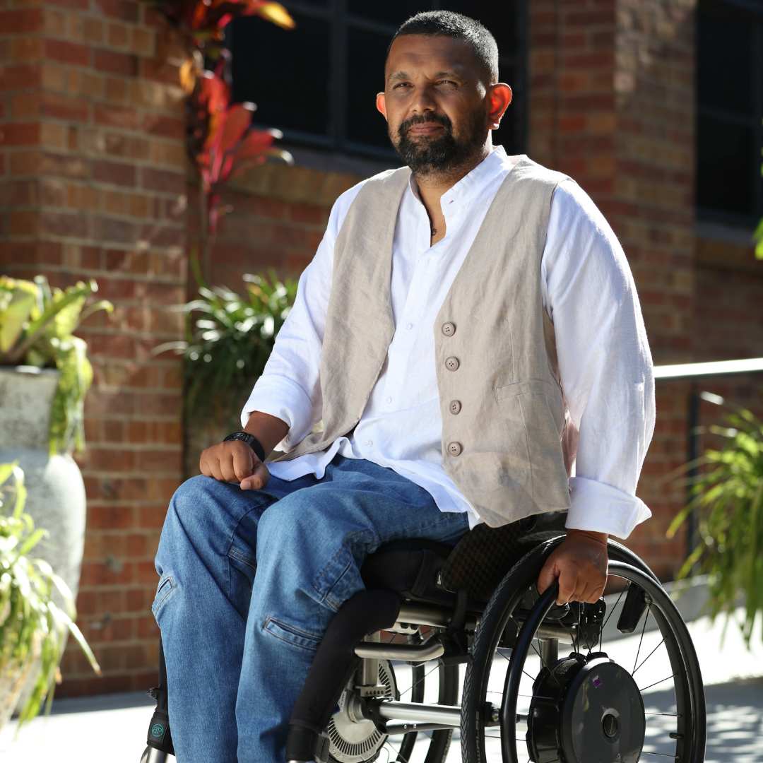 Sam Universal Linen Shirt shown outdoors against brick wall backdrop, styled with beige vest and blue jeans, demonstrating adaptive fashion design with wheelchair-friendly features in natural sunlight and garden setting.