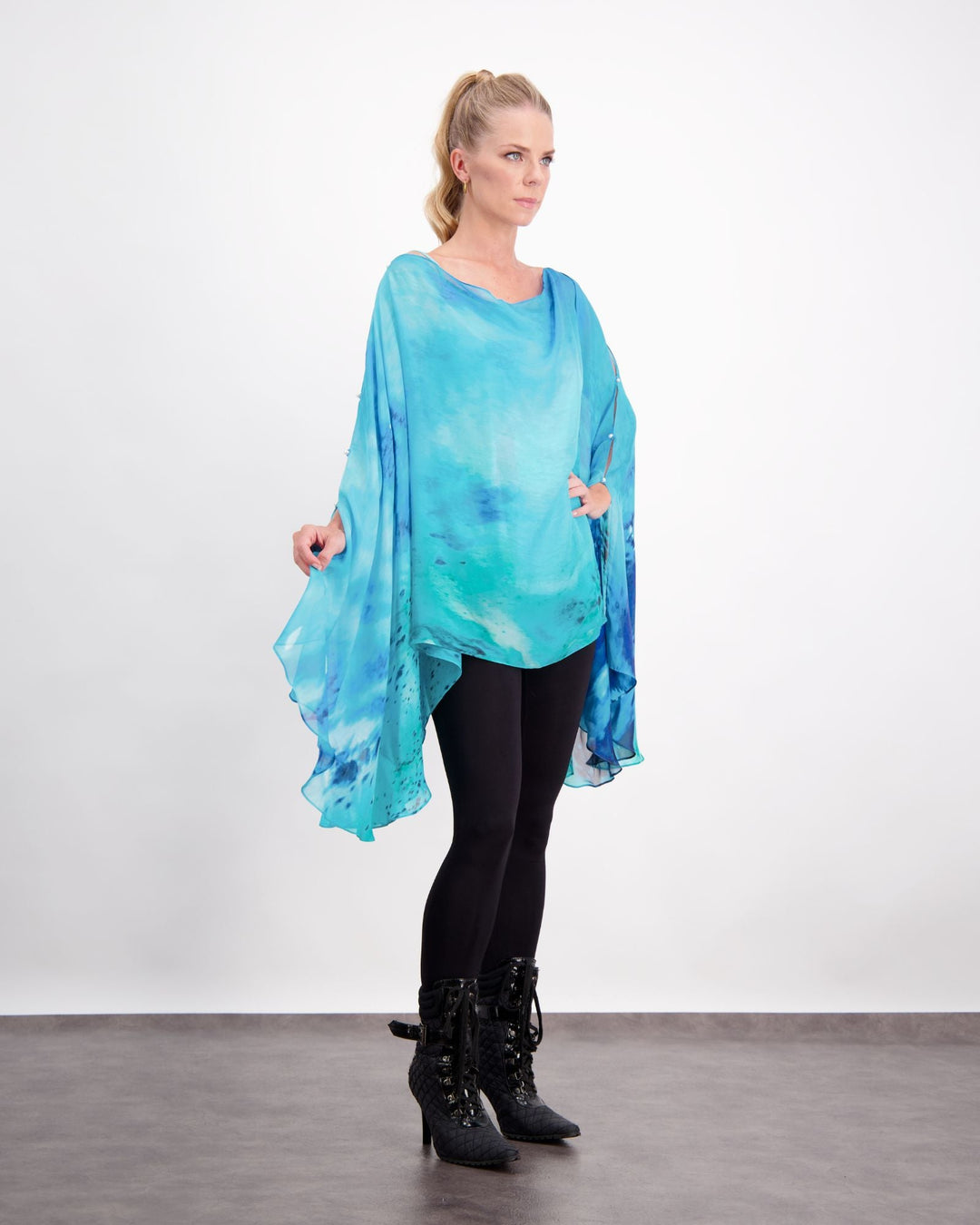 Blonde standing female is wearing a silk, aqua blue print kaftan with small pearl buttons, black leggings and black boots. Christina Stephens Adaptive Clothing Australia. 
