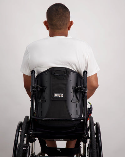 A male in a wheelchair with dark skin tone and dark hair is wearing a white bamboo a-line t-shirt with three small buttons at the neck and brightly coloured board shorts. Christina Stephens Australian Adaptive Clothing.