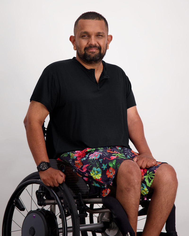 Male in a wheelchair wearing a black t-shirt and colourful boardshorts. Christina Stephens Adaptive Clothing Australia. 