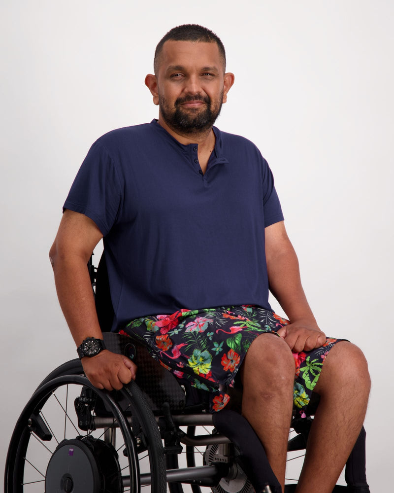 A male in a wheelchair with dark skin tone and dark hair is wearing a navy bamboo a-line t-shirt with three small buttons at the neck and brightly coloured board shorts. Christina Stephens Australian Adaptive Clothing.