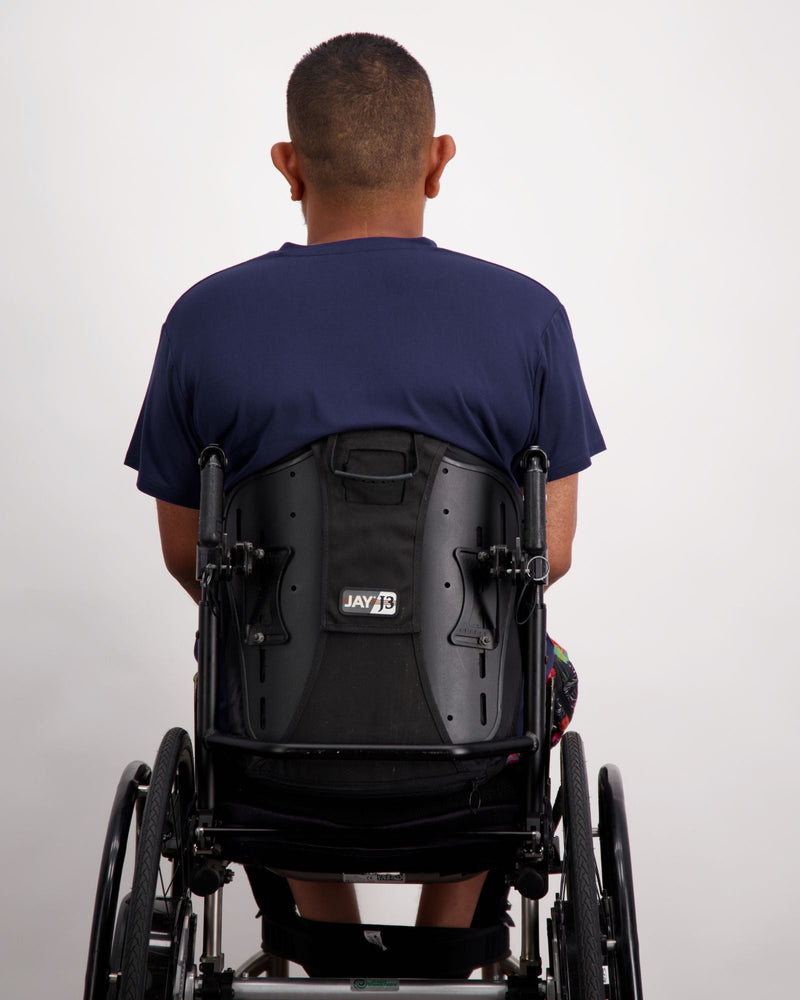 A male in a wheelchair with dark skin tone and dark hair is wearing a navy bamboo a-line t-shirt with three small buttons at the neck and brightly coloured board shorts. Christina Stephens Australian Adaptive Clothing.