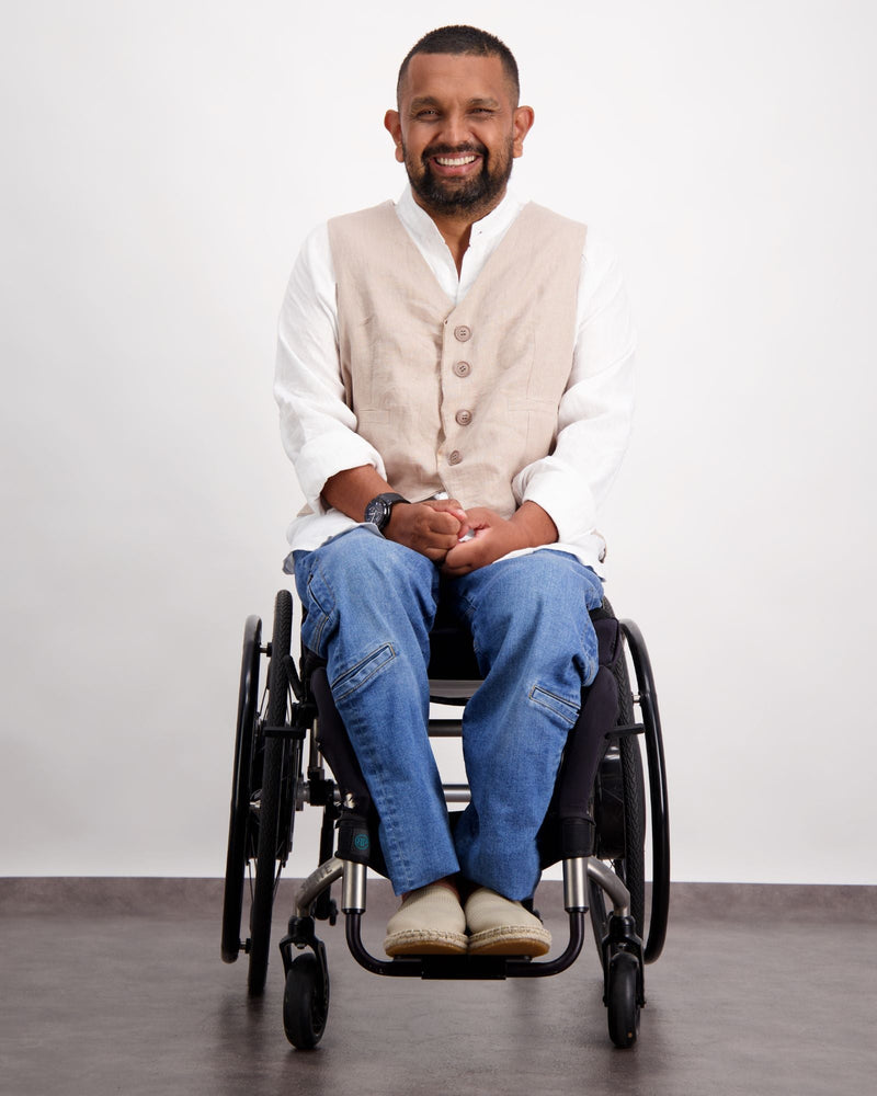 Male with dark skin and dark hair is sitting in a wheelchair wearing a white linen shirt, sand coloured linen vest, blue jeans and sand coloured loafer shoes. Christina Stephens Adaptive Clothing Australia. 