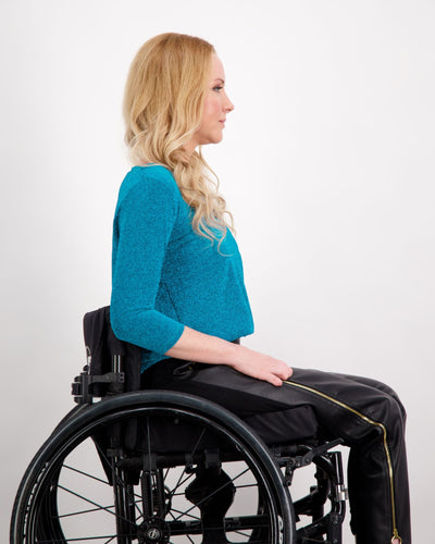 Blonde female sitting in a manual wheelchair is wearing a blue 3/4 sleeve, V-neck glitter top and black vegan leather pants. Christina Stephens Adaptive Clothing Australia.