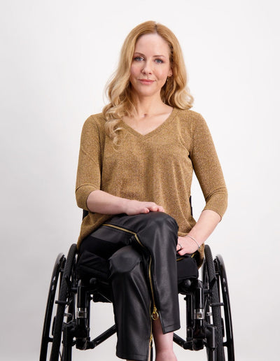 Karni Glitter Top shown in elegant gold metallic fabric with v-neck design and three-quarter sleeves, styled with black leather pants. The adaptive top features a sophisticated leaf-back design, photographed against a clean white background in a wheelchair-friendly composition.