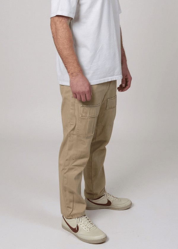 A man visible from the chest down wearing a white t-shirt and beige pants standing against a plain background