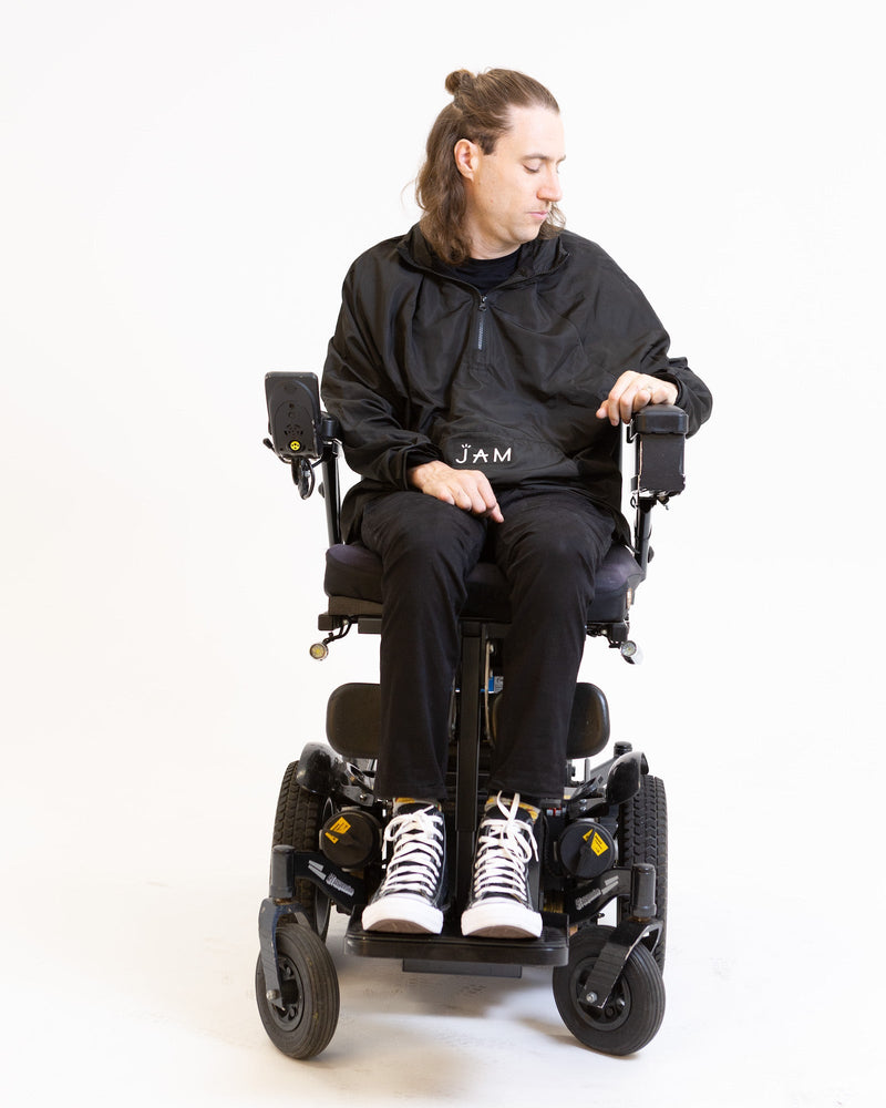 A model seated in his wheelchair in front of a white background. He is wearing a black jacket with black chinos and sneakers. 