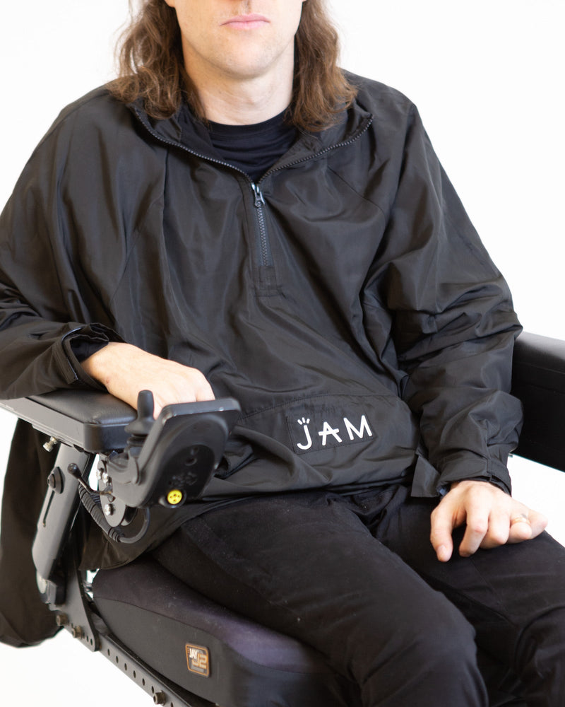 A close up image of a model seated in his wheelchair in front of a white background. He is wearing a black windbreaker jacket with black chinos. 