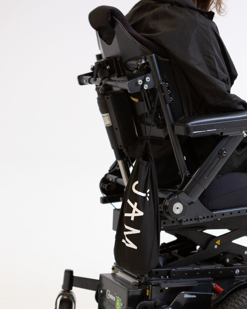 A model seated in his wheelchair visible fro the shoulders down, facing side on to the camera. There is a black drawstring bag with the word "JAM" written on it in white hanging from his wheelchair. 