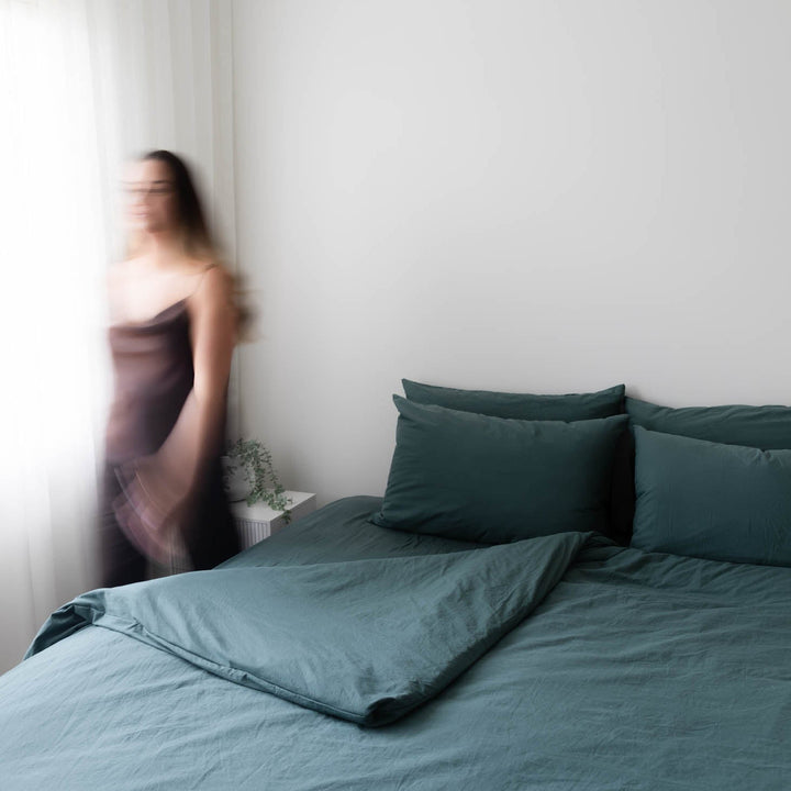 Bed with teal bedding and pillows in a room with a blurred person in the background.