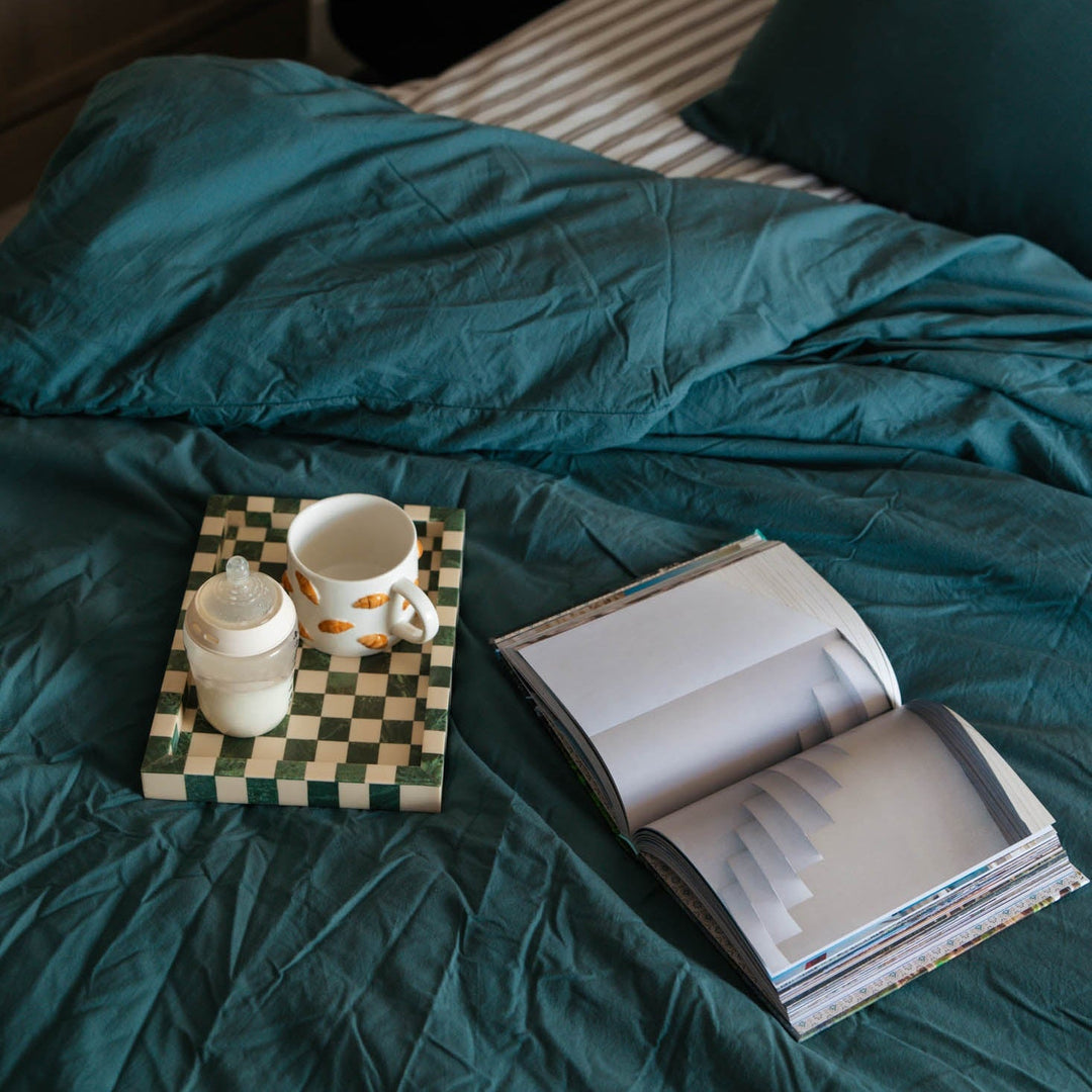 Teal bed with a checkered tray, white mug, and open book on a wooden floor.