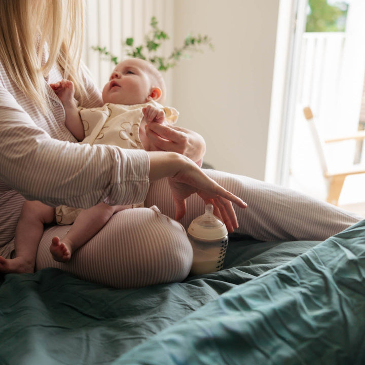 Woman holding a baby and a bottle on a bed in a softly lit room.