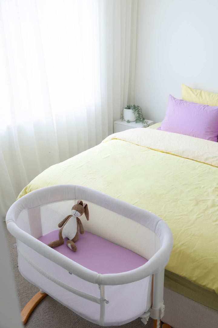 Baby bassinet next to a bed with yellow bedding and purple pillows in a bedroom setting.