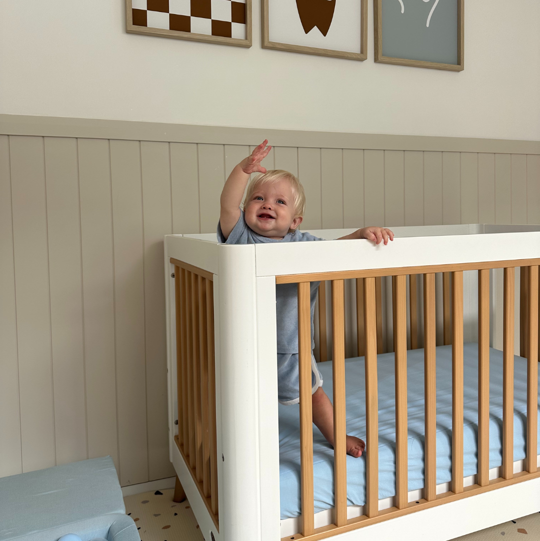 Child in a crib with wooden sides in a room with framed pictures on the wall.