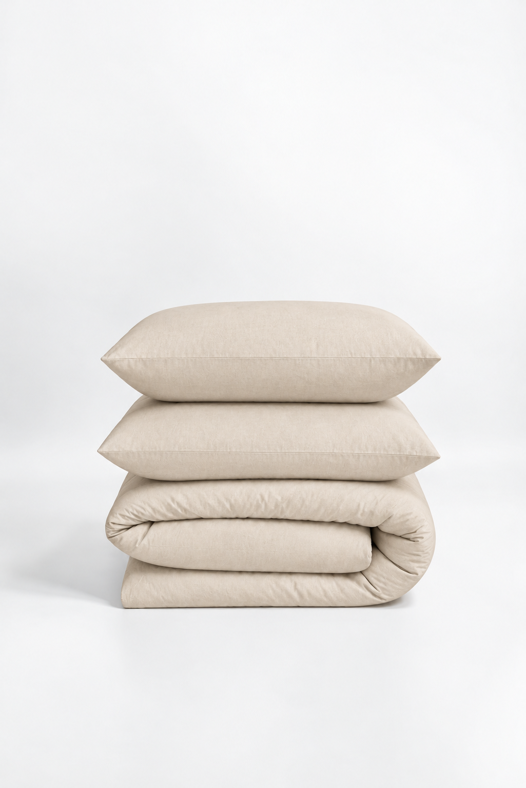 A stack of three light beige bedding items, including two pillows and a folded duvet cover, photographed on a seamless white studio background.