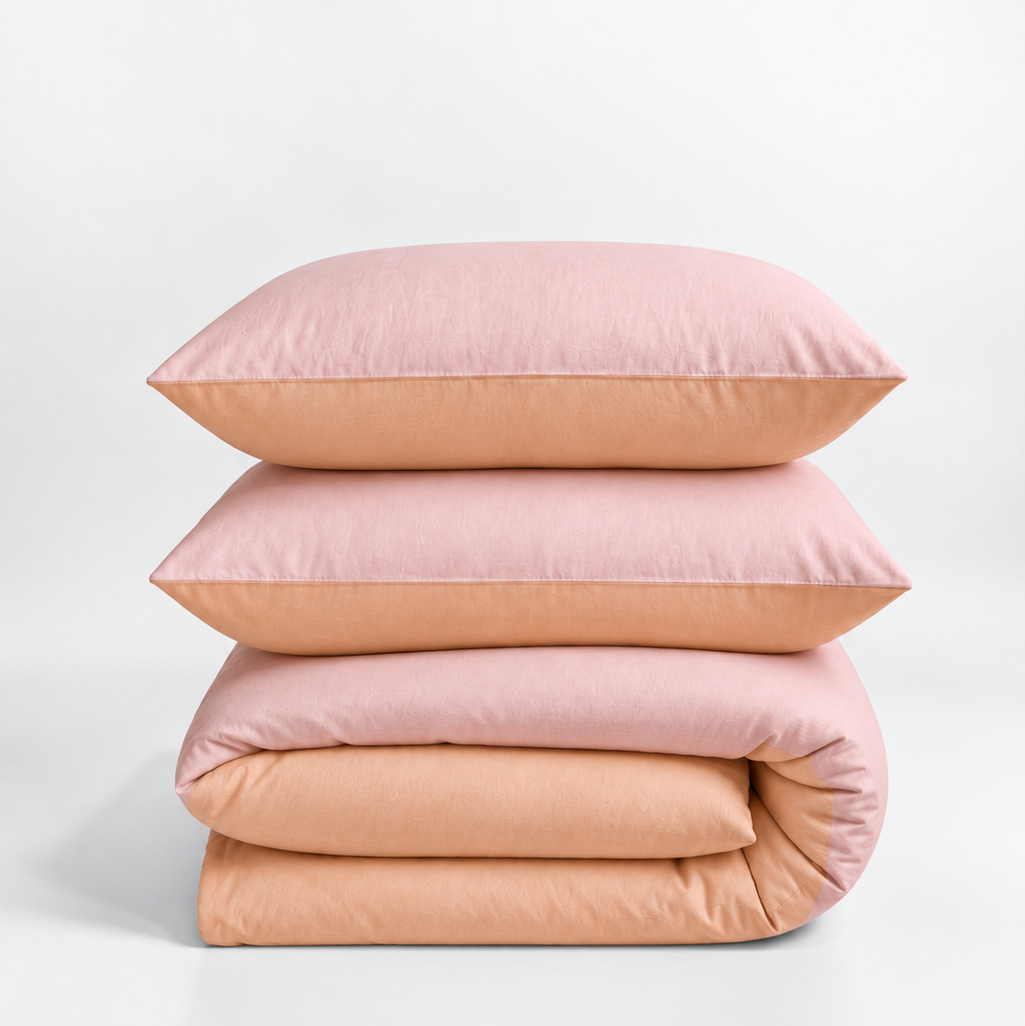 A stack of bedding set including two pink pillows, a folded pink duvet cover, and matching folded sheets in a soft peach color, photographed on a clean white studio background.