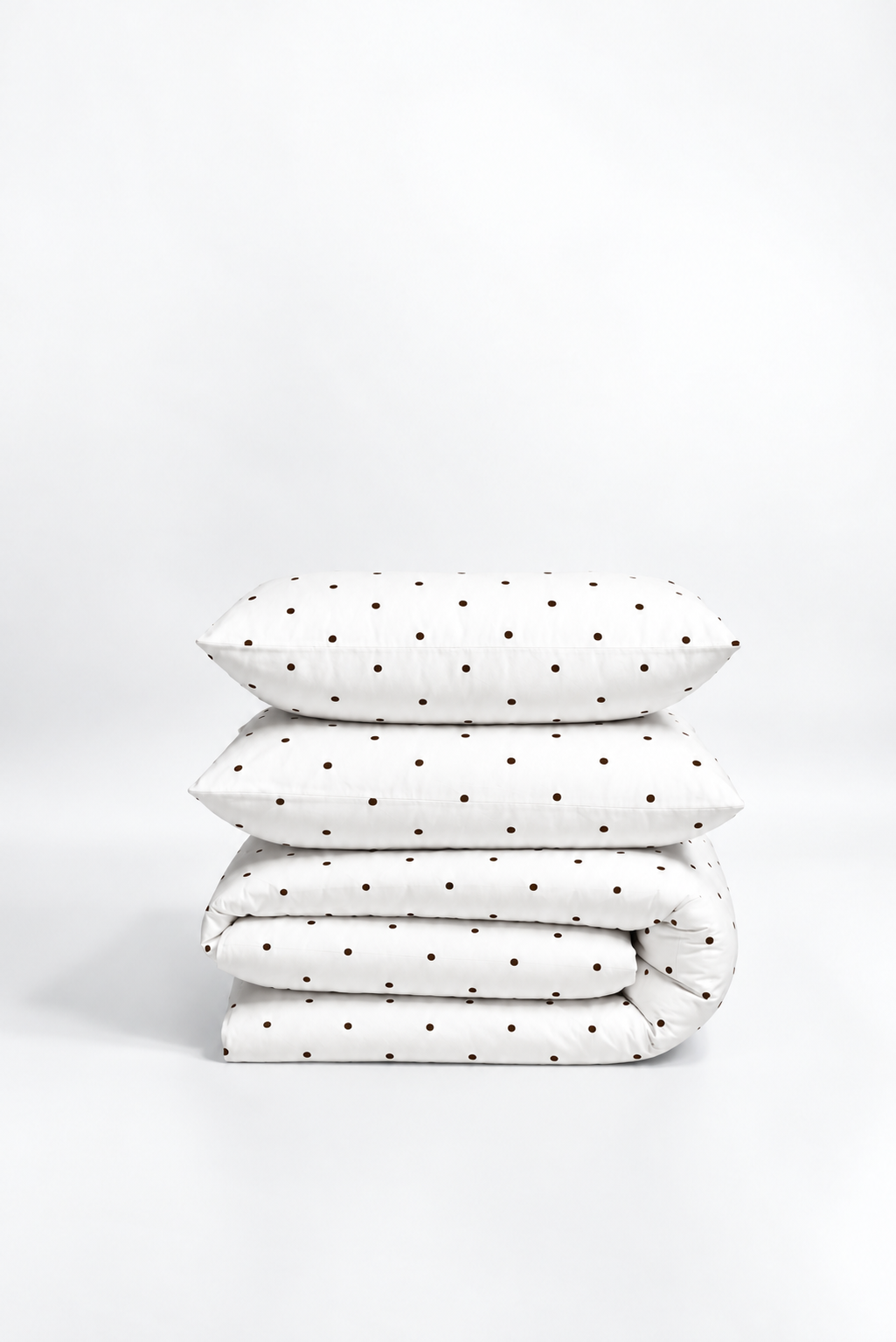 A stack of four white bedding items, including two pillows and two folded blankets, decorated with a small brown polka dot pattern, set against a seamless white studio background.
