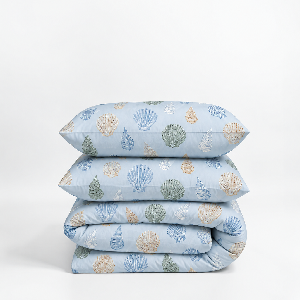 A stack of four light blue bedding items, including two pillows and two folded blankets, printed with a pattern of seashells in blue, green, and beige.