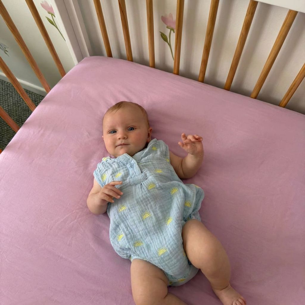 Baby lying on a pink mattress in a crib with a floral patterned headboard.