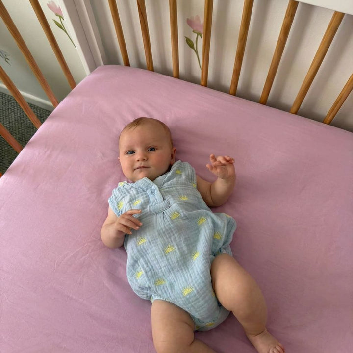 Baby lying on a pink mattress in a crib with a floral patterned headboard.