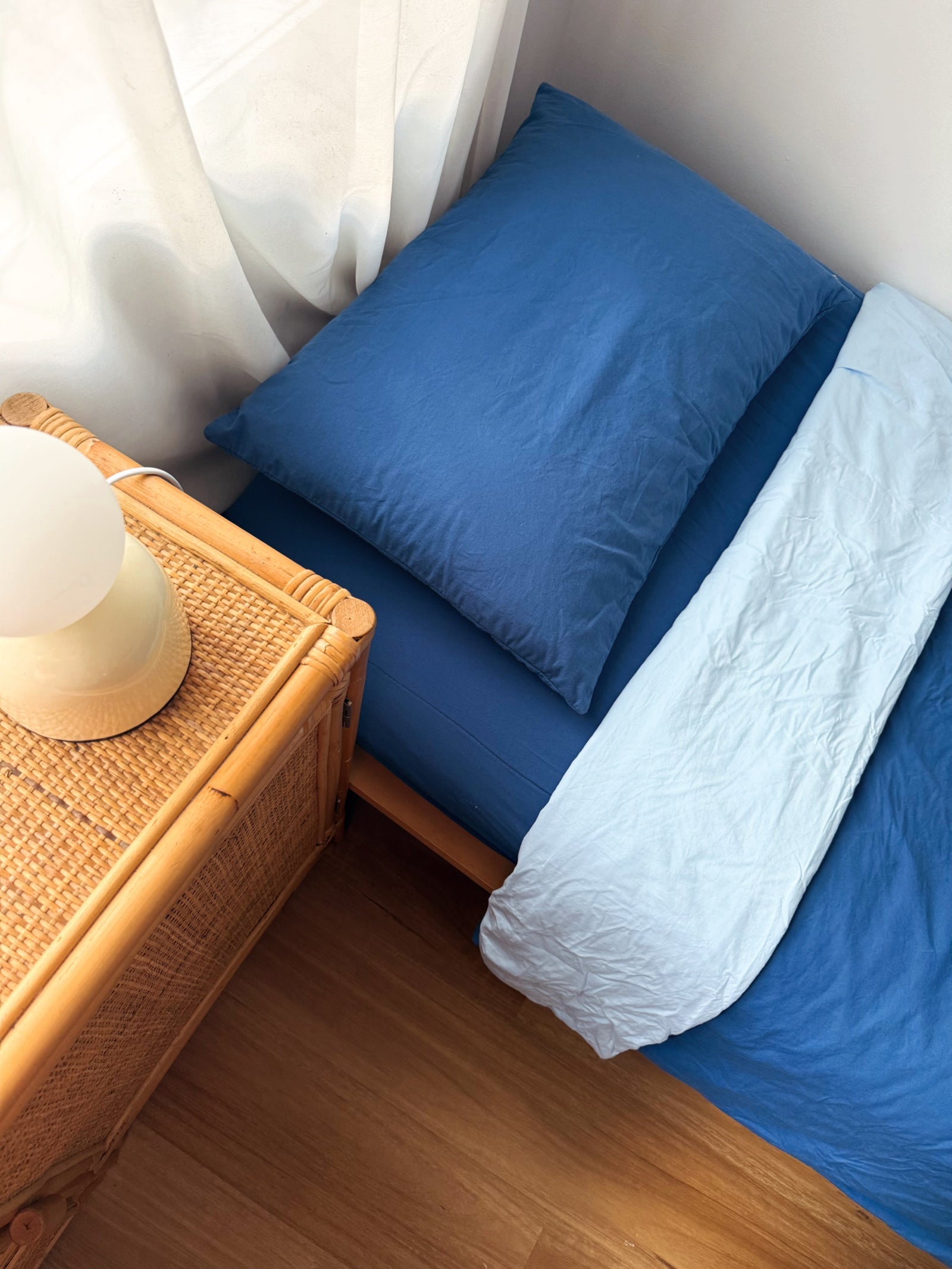 Blue bed with white blanket and blue pillow next to a wooden side table with a lamp.