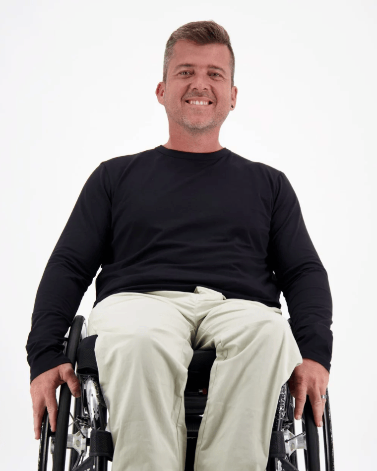 Mens Long Sleeve Leaf Back T-Shirt shown on a person seated in a wheelchair, wearing khaki pants against a white background. The black t-shirt features a classic crew neck design with long sleeves, demonstrating comfortable casual wear that&
