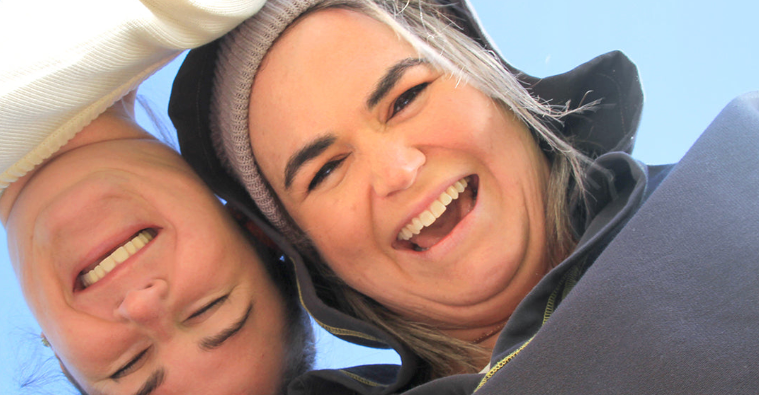 Two people smiling and looking up at the camera with a clear blue sky in the background.
