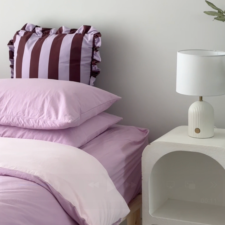 Bedroom with purple bedding and a striped pillow on a bed, next to a white nightstand with a lamp.