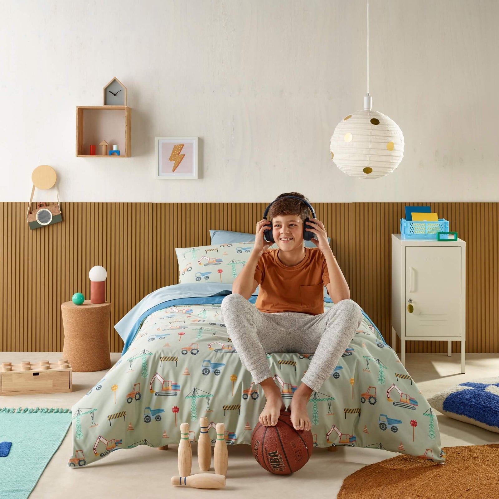 Boy listening to headphones on bed with construction print waterproof bedding and basketball.