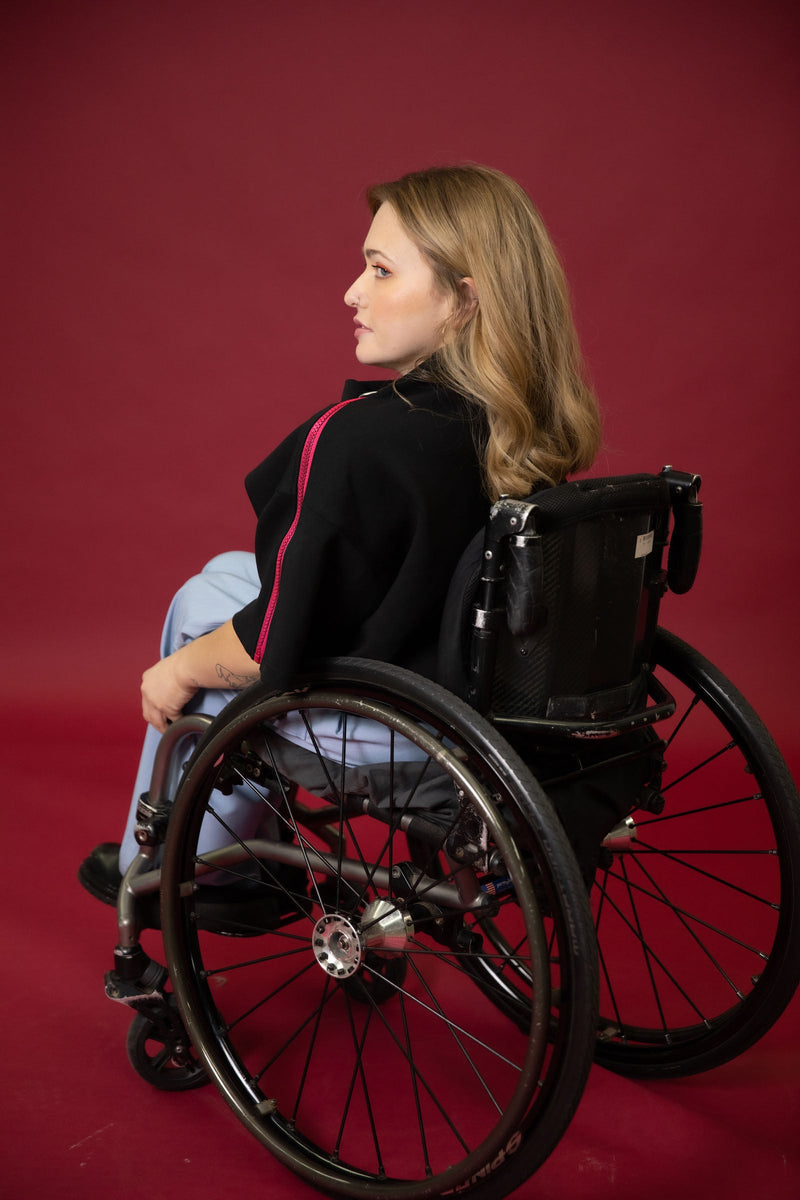 A model seated in her wheelchair facing away from the camera in front of a maroon background wearing a black cropped top with light blue wide leg pants and black shoes 