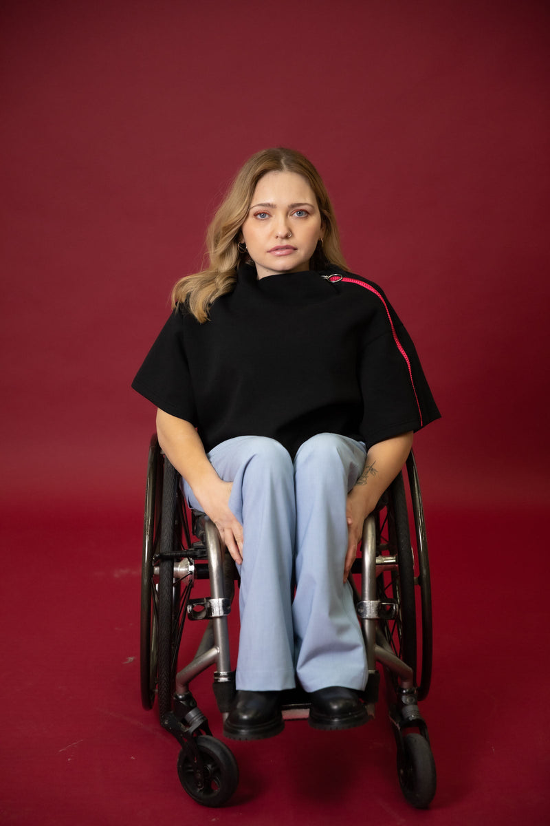 A model seated in her wheelchair in front of a maroon background wearing a black cropped top with light blue wide leg pants and black shoes 