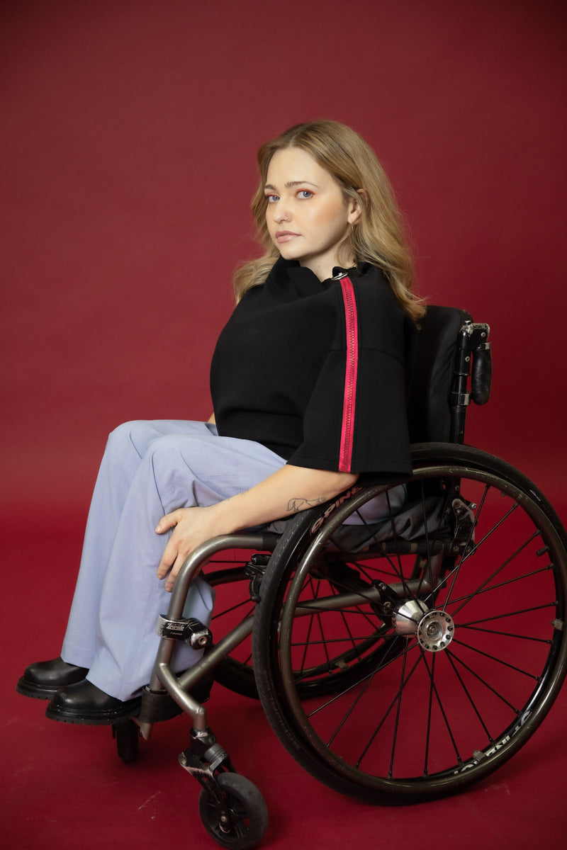 A model seated in her wheelchair side on in front of a maroon background wearing a black cropped top with light blue wide leg pants and black shoes 