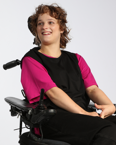 Wicky Smart-Tech Vest shown on a wheelchair user wearing a bright pink shirt underneath the black vest, demonstrating adaptive fashion functionality and comfort. The vest features a sleek, modern design against a light gray background.