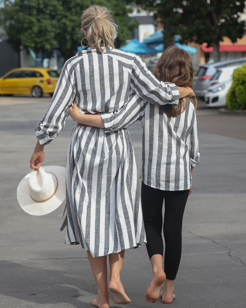 Sapphire Magnetic Shirt Dress shown in casual outdoor setting, featuring elegant black and white vertical stripes, flowing midi length design, with relaxed long sleeves. Summer hat accessory visible, demonstrating the dress&