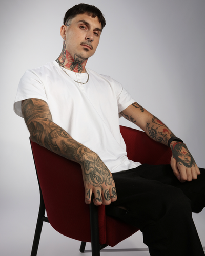 The Nick A-Line Tee shown on a model seated in a burgundy chair against a gray background, displaying the adaptive tee's comfortable fit. Model wears white t-shirt with dark pants, demonstrating the garment's modern silhouette and SpillFree fabric technology.