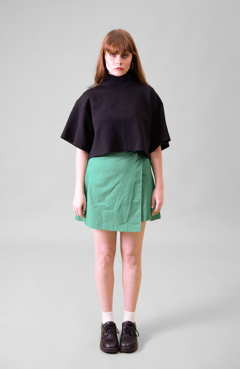 A model standing in front of a grey background wearing a black cropped top and green wrap skirt with black shoes and white socks 