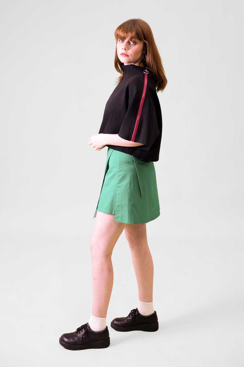 A model standing sideways in front of a grey background facing backwards towards the wall wearing a black cropped top and green wrap skirt with black shoes and white socks 